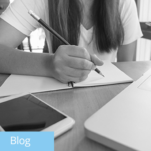 img text: Blog img des: A person sitting at a table writing in a notebook with a phone and laptop nearby